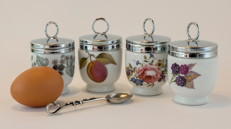 Four vintage egg coddlers next to a brown egg and metal spoon