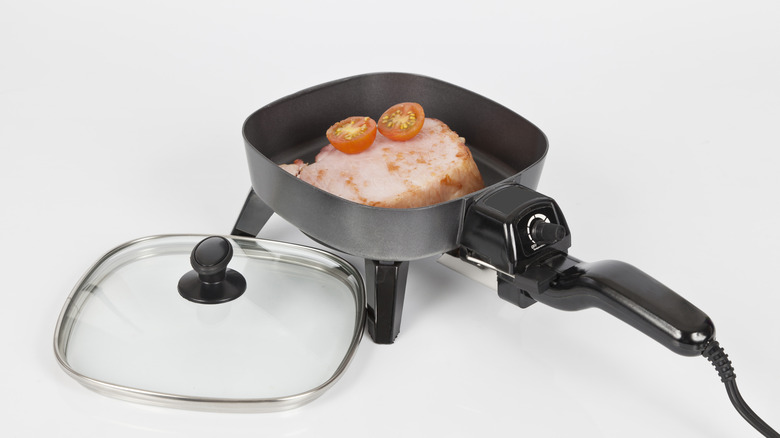 A vintage electric skillet plugged in with food inside and lid next to it
