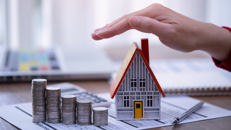 A hand hovering over a miniature house and a pile of coins, sitting on insurance paperwork.