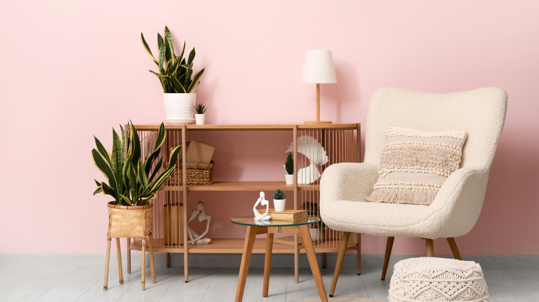 A light pink room with a white chair, wood sideboard, and plants