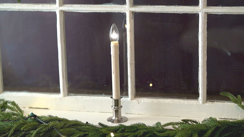 Single battery-powered candle sitting on a windowsill with lighted garland below it