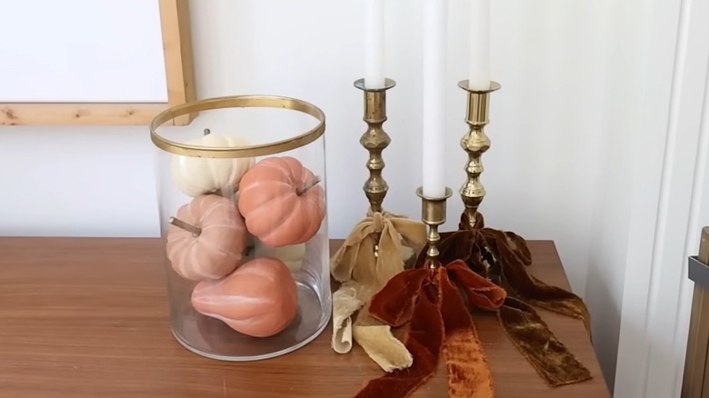 Artificial pumpkins in a glass vase near three candlesticks on a wood table