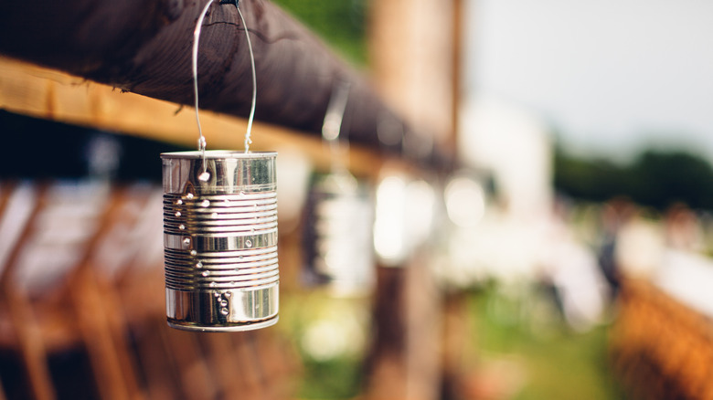 A tin can with holes pierced in it and a candle inside hangs from a fence line from a clothes hanger wire hook.