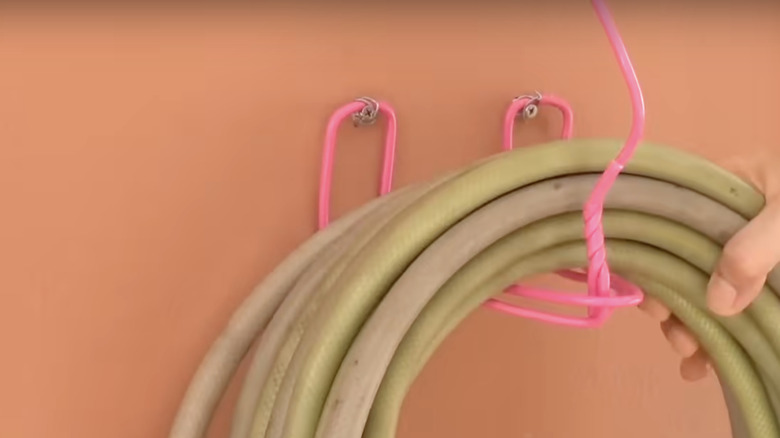 A person loops a garden hose over a wall-mounted hook made from a wire clothes hanger.