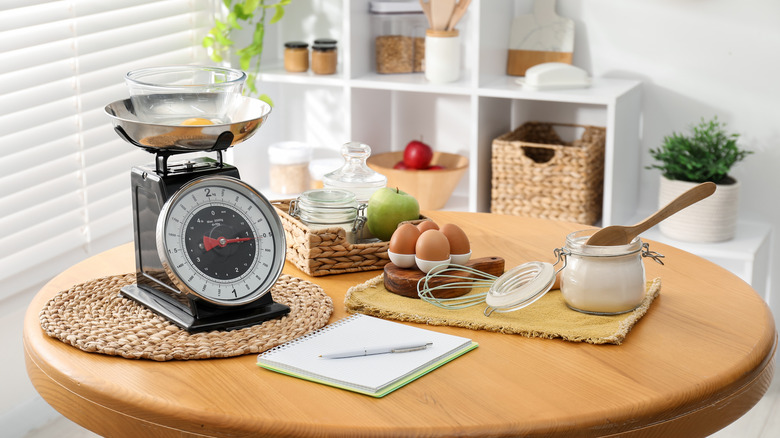 A kitchen table laden baking ingredients, old-school weighing scales, and a handheld whisk