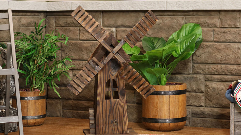 A small wooden windmill on a wooden floor with a brick wall and potted plants in the backgroun