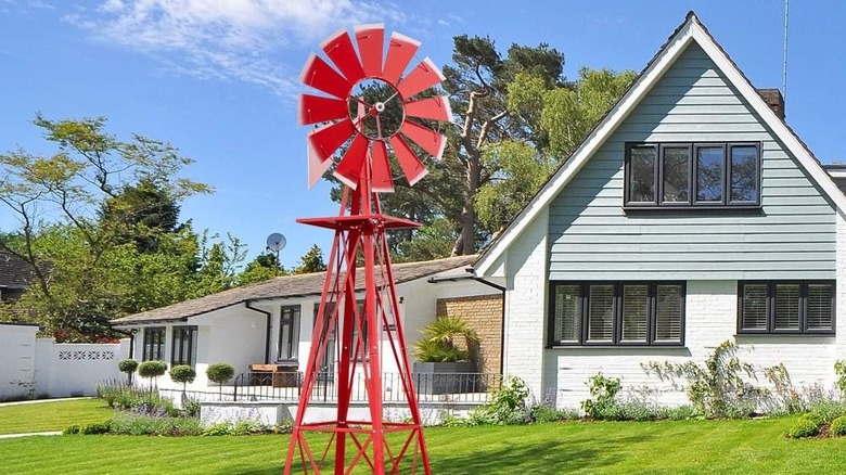 A tall red windmill with small stripes of white on the blades standing on a green lawn in front of a house