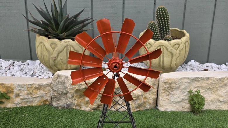 A bright orange windmill on dark metal legs on grass with rocks and succulents behind