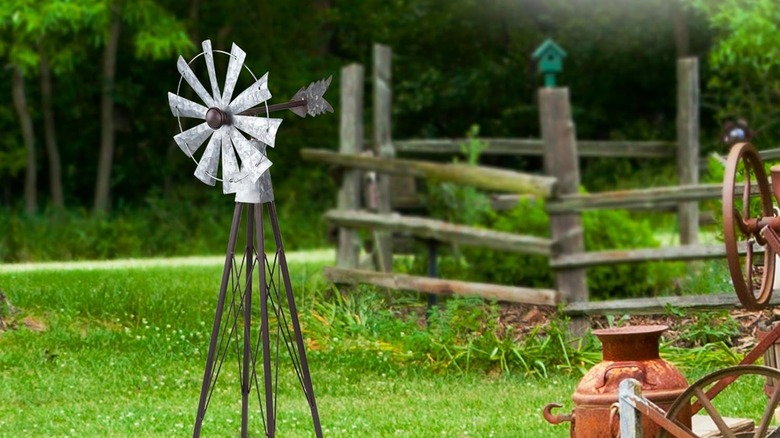 A silver metal windmill with a darker metal stand and arrow fin on a green field with an old wooden fence and other antique pieces nearby