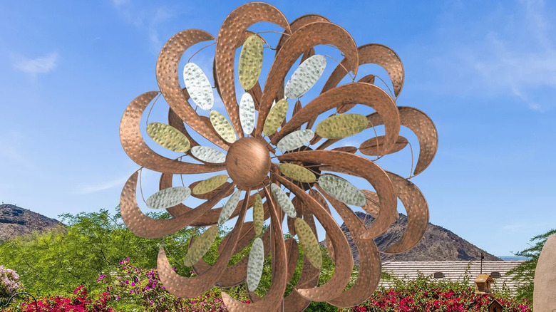A copper, flower-like windmill with trees, flowers, and clouds in the background