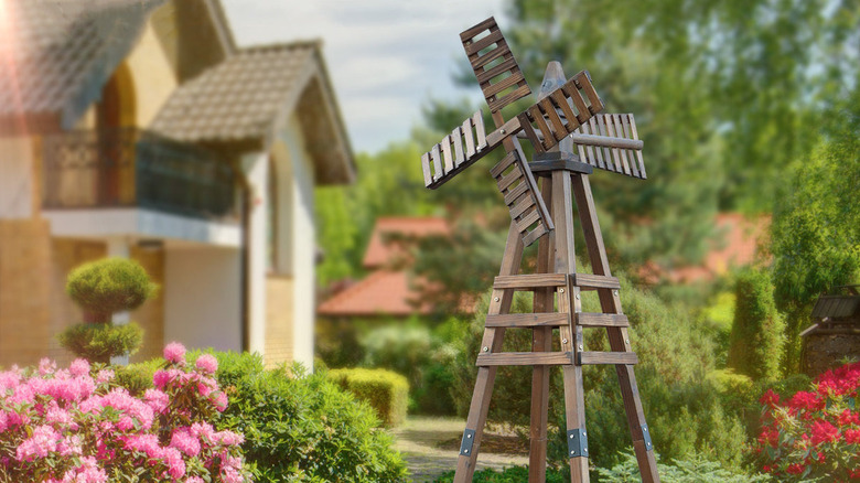 A wooden windmill surrounded by greenery with a house in the background