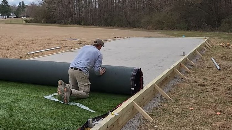 A man rolls turf on top of a raised gravel base for a batting cage.