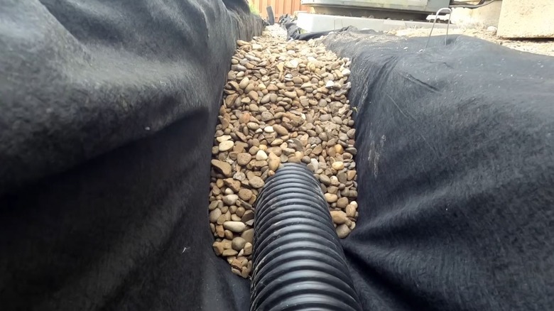 A French drain being covered with gravel in a ditch lined with landscape fabric.