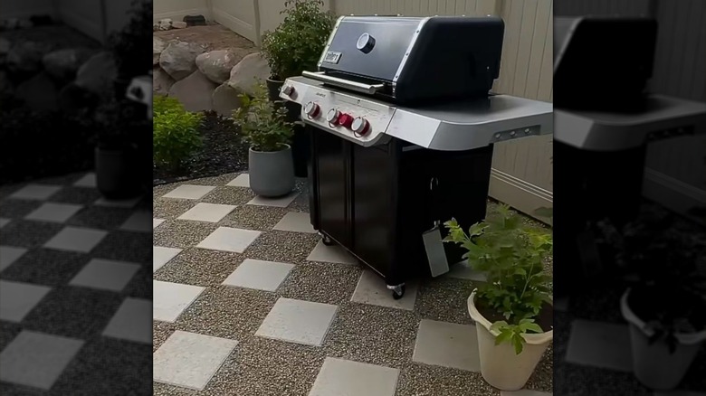 An outdoor grill rests on a gravel and paver stone patio surrounded by potted plants.