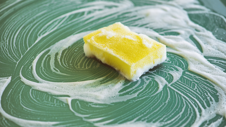 Kitchen sponge on green work surface with cleaning product foamed up around it in swirls.
