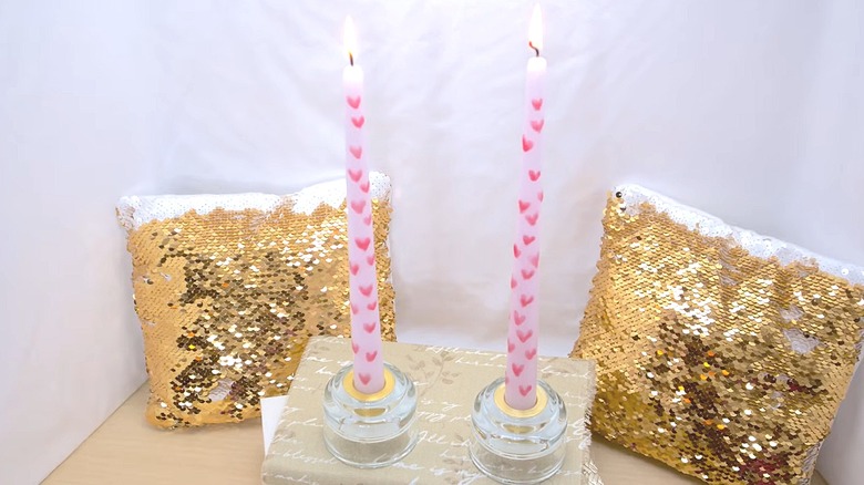 Two taper candles with hand-painted hearts on them with gold sequin pillows in the background