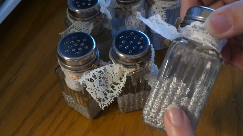 A hand holds a salt and pepper shaker filled with silver glitter and adorned with lace ribbon in front of similar shakers on a wooden table.