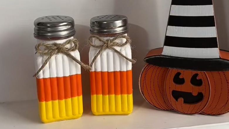 Two shakers painted like candy corn on a white shelf with a pumpkin decoration
