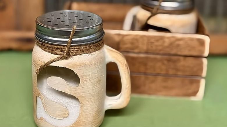 A salt shaker with a peekaboo S painted on it and a pepper shaker in a wooden box behind it.