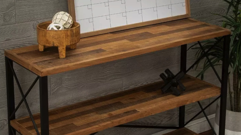 A standing wooden shelving unit with home decor items and a calendar on it near a potted plant