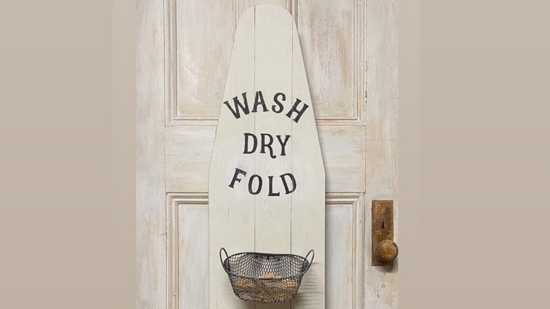 Ironing board painted white with "Wash Dry Fold" painted on it and a basket attached
