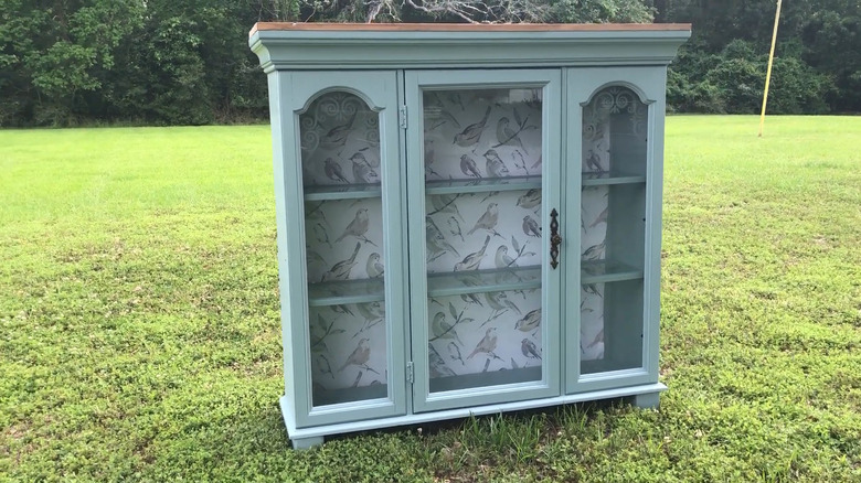 Light green hutch with bird wallpaper and a stained top repurposed into a glass-front storage cabinet