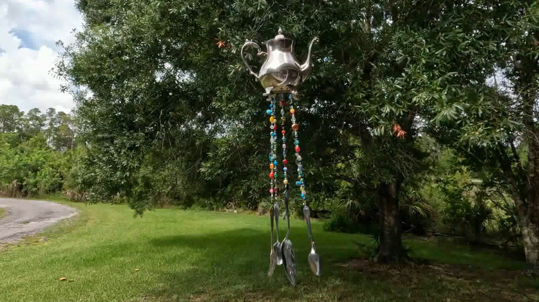 Metal tea pot with strings of beads and spoons on the end creating a DIY wind chime hanging in a tree