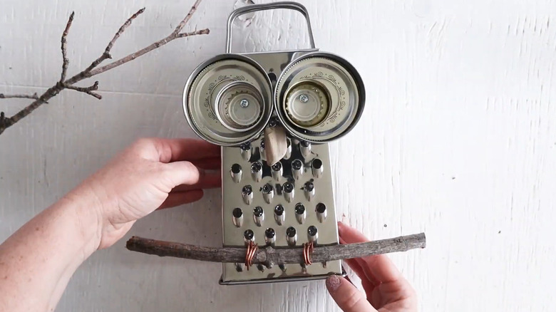 Person picking up an owl decoration made with a cheese grater and lids for the eyes with a stick at the bottom