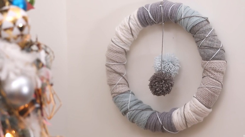 Wreath form covered with sections of sweaters with yarn pompoms hanging in the middle