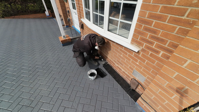 A man paints a gray paver driveway with a border of black paint.