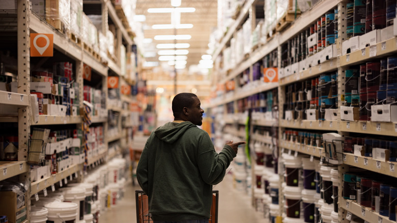 A person pushing a shopping cart peruses the shelves of a hardware store full of paint cans.