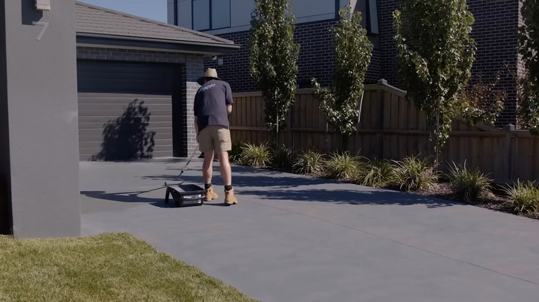 A person uses a roller and tray to paint a concrete driveway with gray concrete paint.
