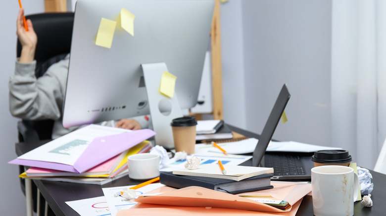 A messy desk with sticky notes, pencils, crumpled paper, andpaper clutter all over it, and a person partially hidden behind a computer screen