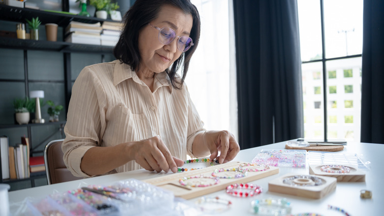 An older woman making bracelets out of multiple beads on a table inside a home