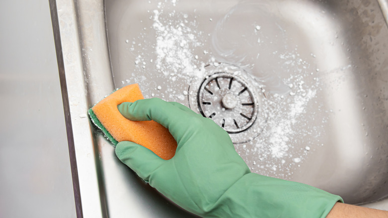 Person wearing gloves and cleaning kitchen sink with an orange sponge