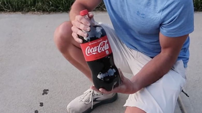 A man holds a bottle of Coca-cola while squatting on a concrete driveway.