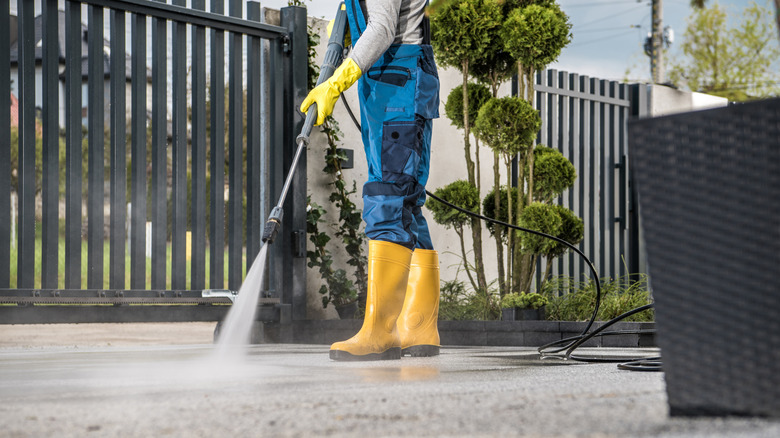 A person in bibs and rubber boots using a powerwasher on a driveway with plants and a fence.