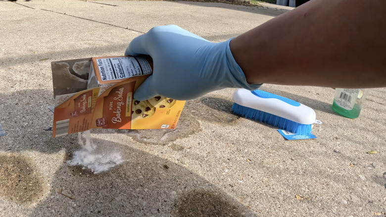 White powder in a small glass jar labeled baking soda with a small spoon with the powder on it sitting on a concrete surface.