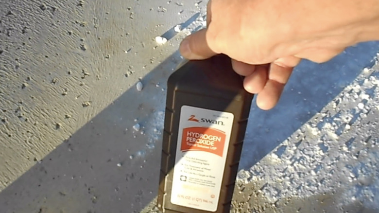 A person holds a bottle of hydrogen peroxide above a concrete driveway sprinkled with baking soda.