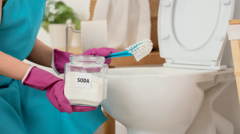 Someone kneeling in front of toilet wearing rubber gloves and holding scrub brush and jar of baking soda
