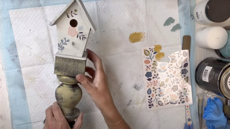 A crafter glues a wooden birdhouse to the top of a repurposed and painted deck railing spindle.