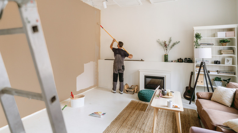 A man uses a roller to paint tall white living room walls beige.