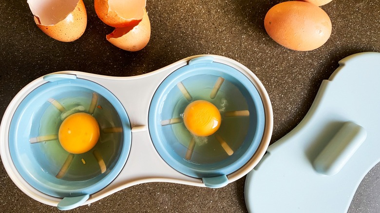 A microwave egg poacher holding two eggs with shells on the countertop.