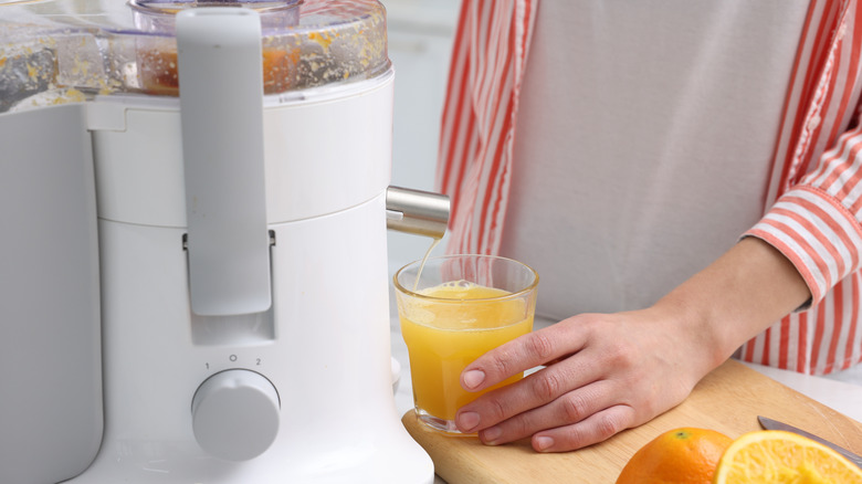 An electric juicer in use with a glass of freshly squeezed orange uice being held by a man in a red and white striped shirt and white t-shirt.