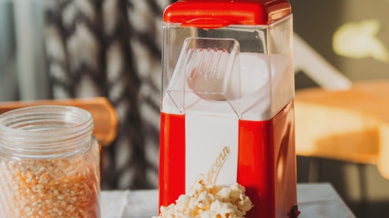 A bright red popcorn maker next to a jar of popcorn.