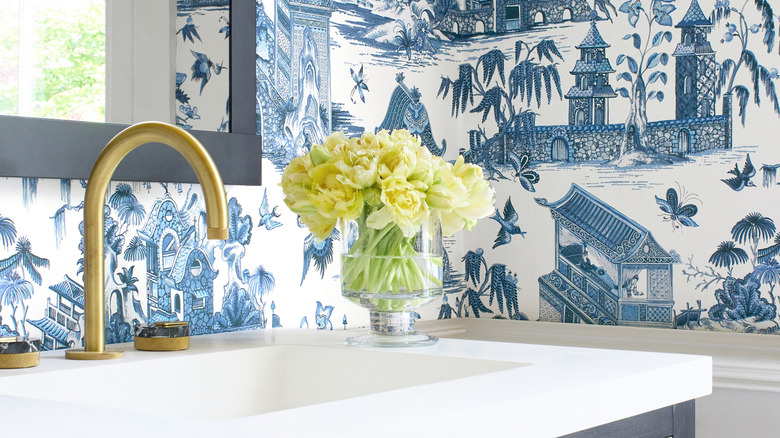 A bathroom sink with a gold tap, fresh flowers in a vase, and stylized Chinese wallpaper.