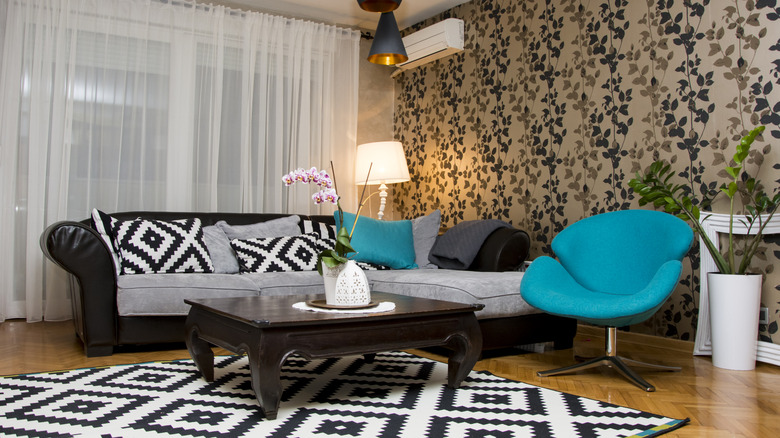 A living room with chaise longue style sofa, teal modern armchair and low coffee table, with a modern black and white geometric rug and floral wallpaper.