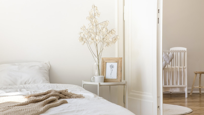 A white bedroom with brown throw. A bedside table contains a small plant, print of the plant in a pine frame and a white cup.