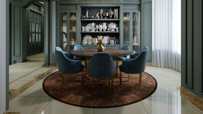 A round dining table on a round brown rug with blue dining chairs spaced around it. There is a large fitted sideboard behind it full of vases and jugs in silver and white.
