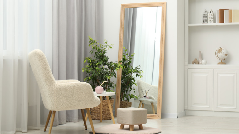 A large, freestanding mirror behind an armchair and potted plant next to a window.